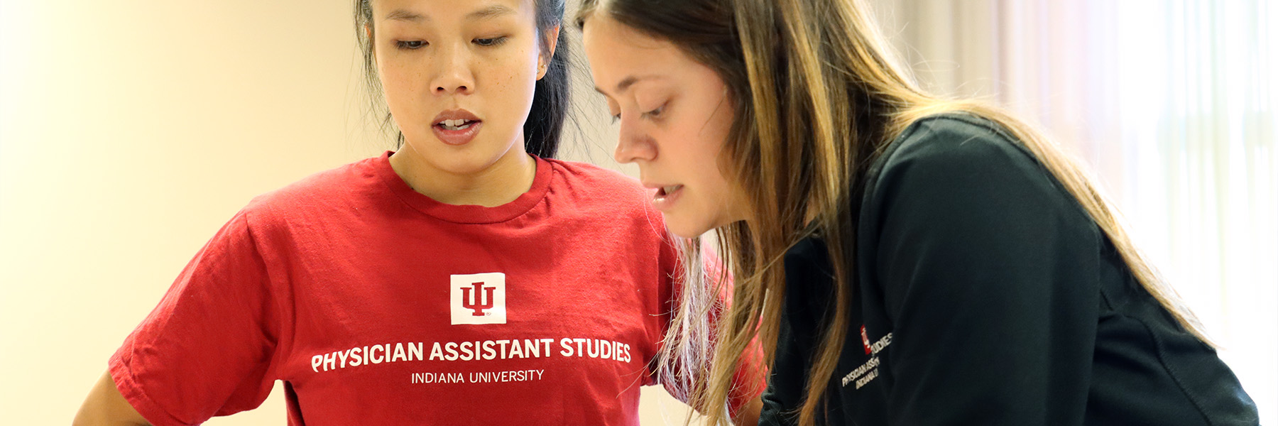 Two students in IU Physicians Assistant tee shirts work lean toward each other talking