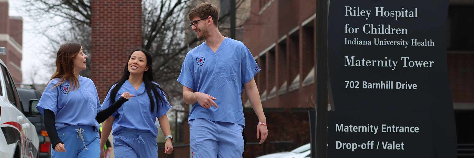 Three students in blue scrubs walk on a sidewalk approaching a sign for Riley Hospital.