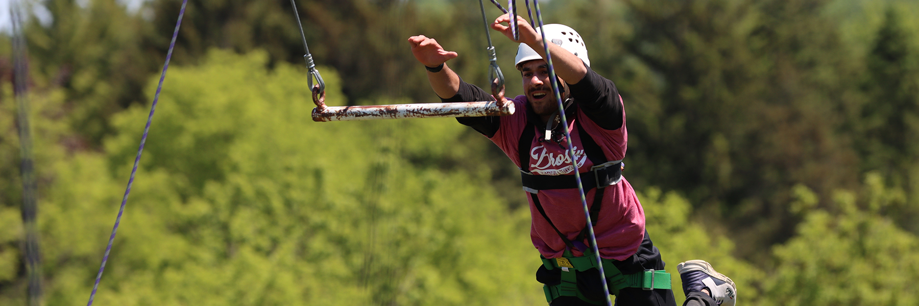 A male in a hemet and strapped into protective gear leaps forward to grab onto a hanging bar at a outdoor adventure course.