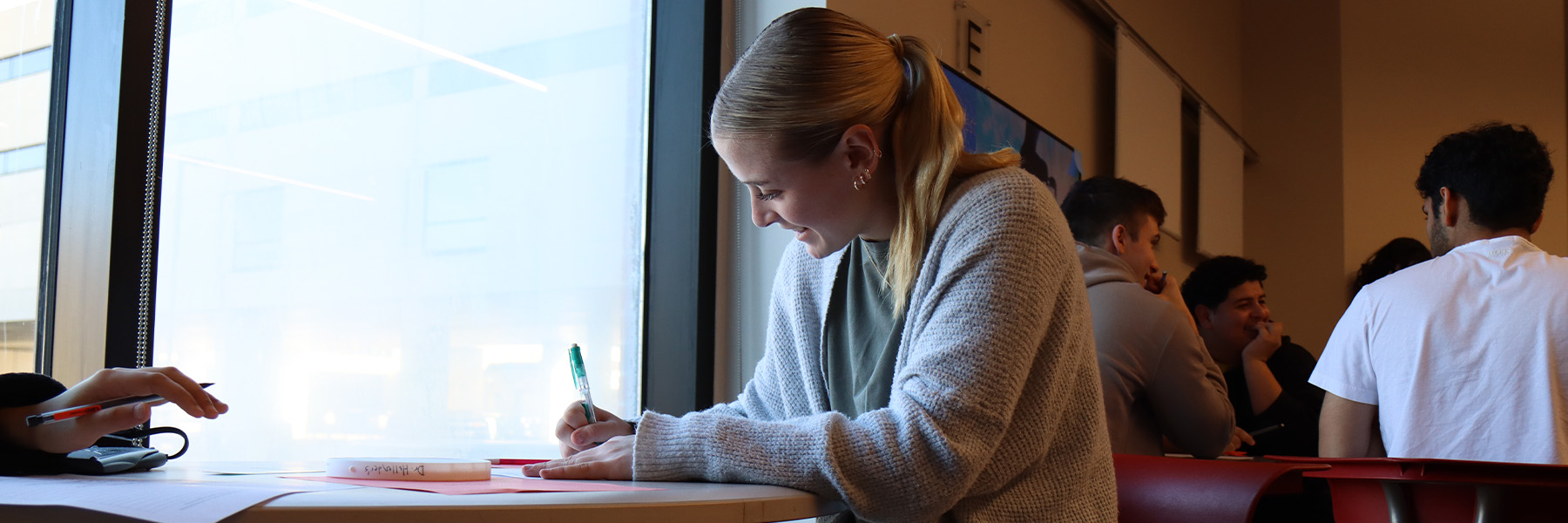A female student writing on a piece of paper at a desk near a bright window; students at another table are seen in the background