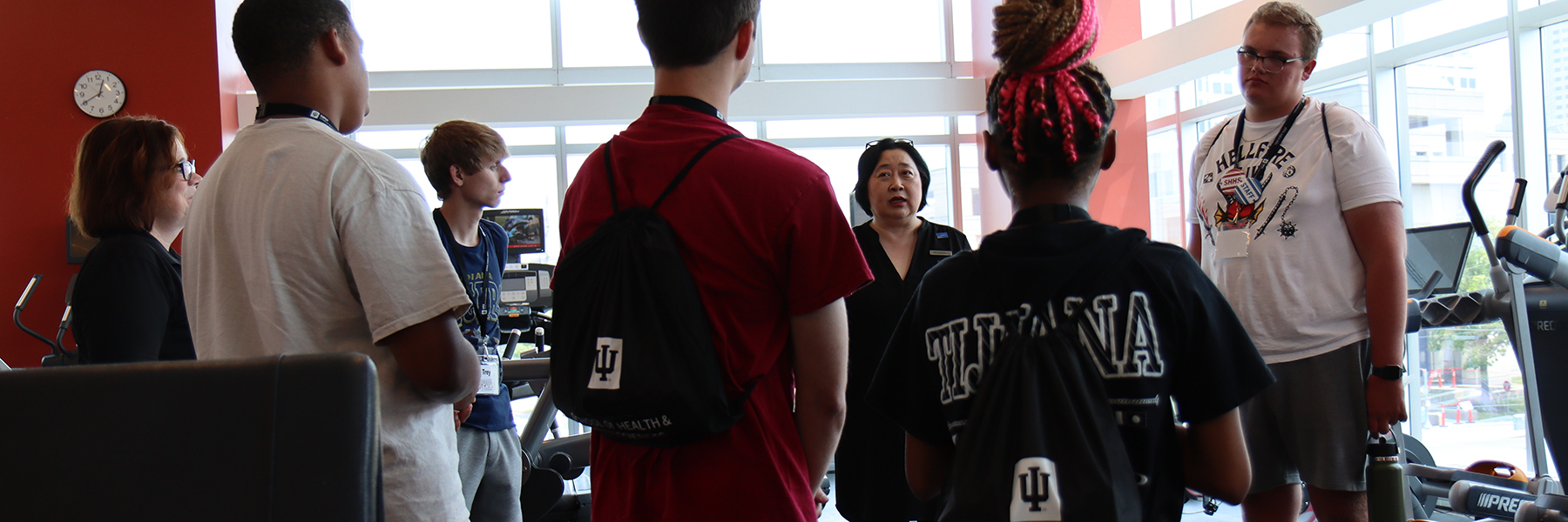 A group of students stand in a semi-circle around a woman speaking to them in a gym space of cardio machines.