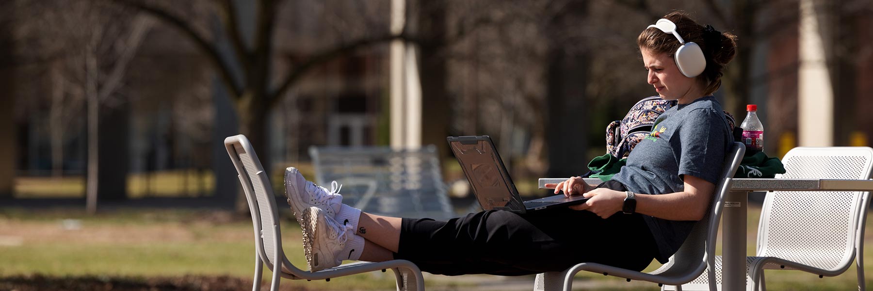 A student studies with a laptop on her lap and headphone on while sitting with her legs up on an outdoor table and chairs