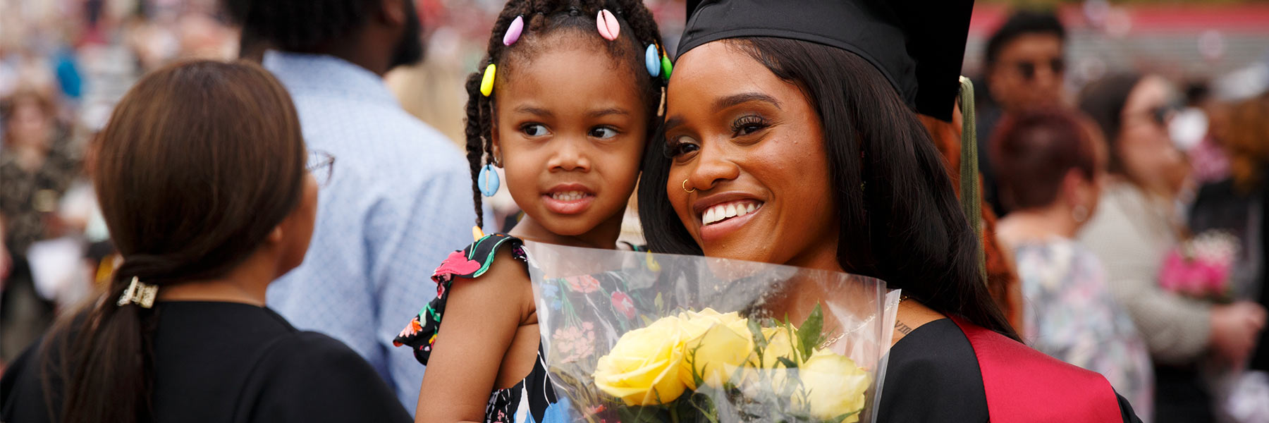 A graduate in cap and gown holds flowers with one arm and a young girl in her other arm; other commencement attendees seen in background